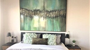 A bedroom with a large abstract painting above the bed, a grey and white bedspread, and two bedside tables with lamps and decorative items.