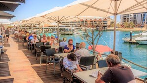 A waterfront outdoor dining area with tables and chairs under large umbrellas, overlooking a marina with boats.