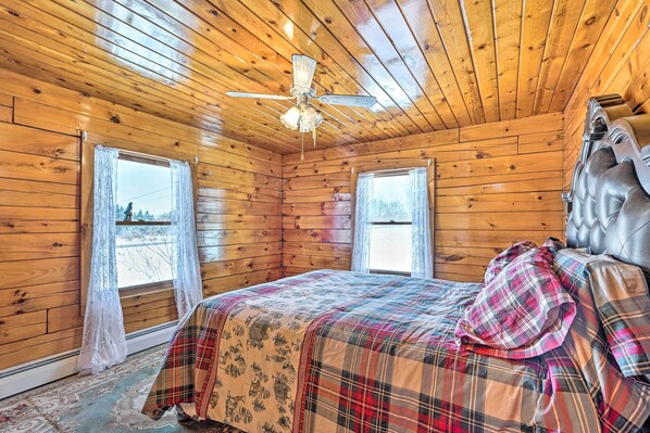Un dormitorio de cabaña de madera con una cama, un ventilador de techo y dos ventanas con cortinas.