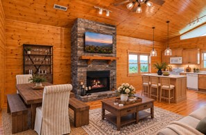 A cozy cabin interior with a stone fireplace, wooden furniture, a dining area, and a kitchen.