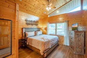 A wooden cabin bedroom with a bed, a nightstand, a dresser, a ceiling fan, and a window with curtains. - Fall DEAL! Hillside Mountain Views & Hot Tub! (Sevierville)