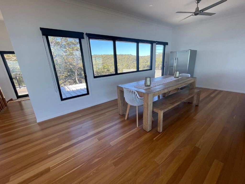 Une pièce spacieuse dotée d’un plancher en bois, d’une table à manger avec des chaises et de grandes fenêtres offrant une vue sur les arbres et les collines.