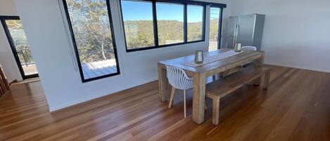 Une pièce spacieuse dotée d’un plancher en bois, d’une table à manger avec des chaises et de grandes fenêtres offrant une vue sur les arbres et les collines.