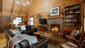 A cozy log cabin interior with a fireplace, leather sofa, wooden chairs, and a bookshelf.
