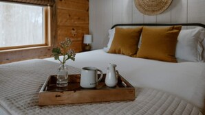 A neatly made bed with white and mustard pillows, a bedside table with a vase of flowers and a tray with a mug and pitcher.