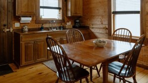 A wooden cabin interior with a dining table, chairs, a kitchen area, and a window.