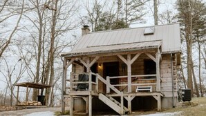 Un chalet rustique doté d’un porche et d’une cheminée, entouré d’arbres et bordé d’un chemin de gravier.