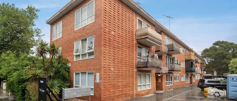 A multi-story residential building with a brick facade, balconies, and a parking area.