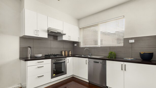 A modern kitchen with white cabinets, a black countertop, stainless steel appliances, and a window with blinds. - Poets Retreat (Elwood)