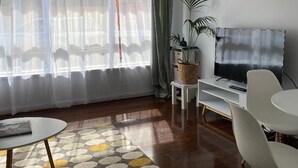 A modern living room with a large window, a flat-screen TV, a dining table, and a potted plant. - Poets Retreat (Elwood)