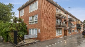 A multi-story brick building with balconies, a parking lot, and a small garden area. - Poets Retreat (Elwood)