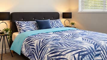A bedroom with a bed, a cityscape painting, a window with blinds, and a bedside table with a lamp.