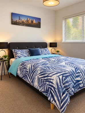 A bedroom with a bed, a cityscape painting, a window with blinds, and a bedside table with a lamp. - Poets Retreat (Elwood)