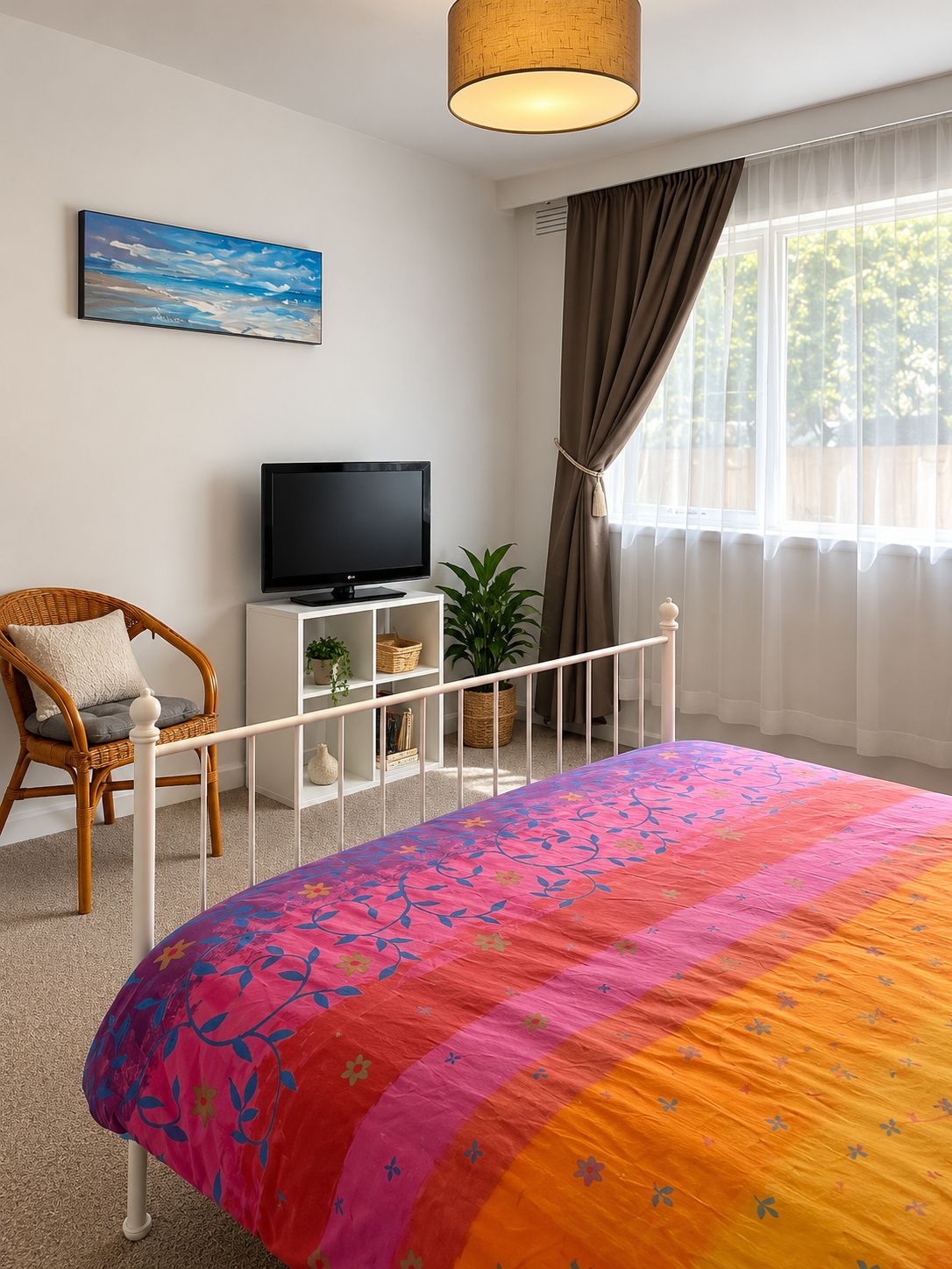 A bedroom with a bed, a television, a wicker chair, a white bedside table, a window with curtains, and a painting on the wall.