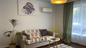 A living room with a sofa, a small table, a potted plant, a colorful rug, and a large window with curtains. - Poets Retreat (Elwood)