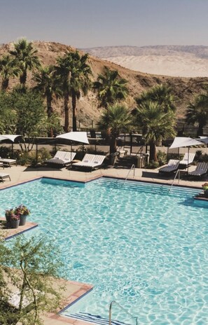 A swimming pool with lounge chairs and umbrellas, surrounded by palm trees and a desert landscape.