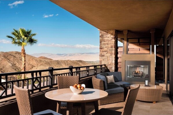 A balcony with wicker furniture, a round table with a bowl of fruit, and a fireplace.