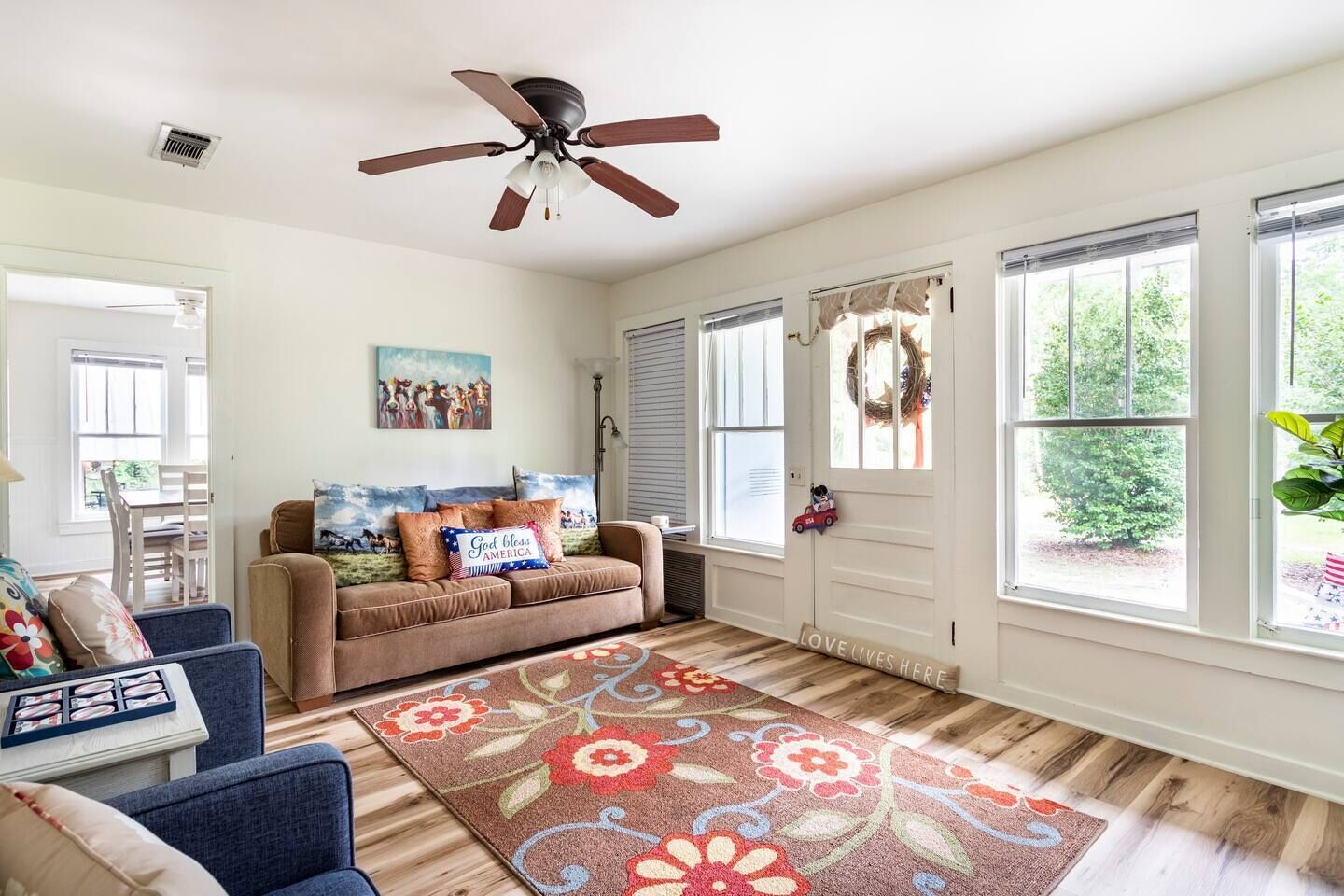 Uma sala de estar com um sofá marrom, uma poltrona azul, um tapete floral e um ventilador de teto.