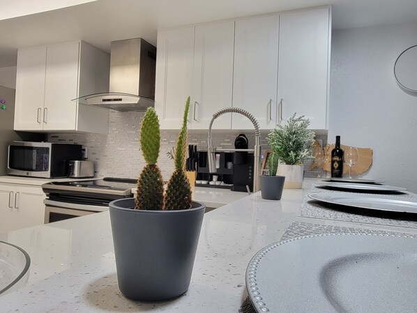 A modern kitchen with white cabinets, a stainless steel range hood, a microwave, and a coffee maker. There are two potted plants on the counter.
