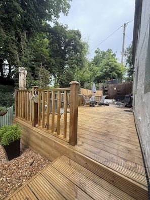 Una terraza de madera con barandilla, rodeada de árboles y un área cercada con muebles de exterior.