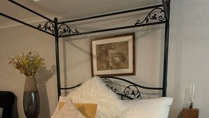A neatly made bed with white linens, a fluffy white throw, and a rolled white towel. There is a framed picture on the wall, a vase with flowers on a nightstand, and a wooden nightstand with drawers. - “Quirky Cottage” 2 double rooms (Dumfries)