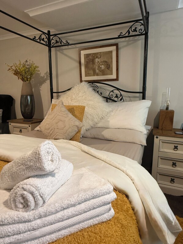 Una cama bien hecha con ropa blanca, un suave plaid blanco y una toalla blanca enrollada. En la pared hay un cuadro enmarco, un florero sobre una mesita de noche y una mesita de noche de madera con cajones.