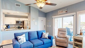 A modern living room with a blue sofa and a beige armchair, a kitchen with white cabinets, and a large window offering a view of the outdoors.