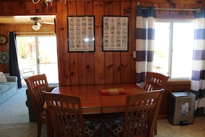 A wooden dining room with a table and chairs, framed pictures on the wall, and a window with curtains.