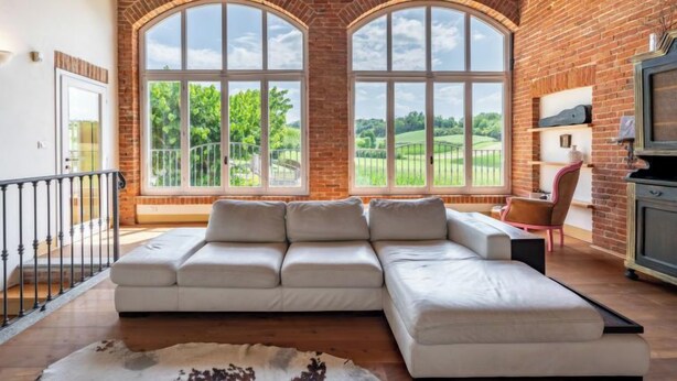 A spacious living room with a large sectional sofa, a rustic rug, and a brick wall with arched windows offering a view of a green landscape. - La Bell'aria by Interhome (Frinco)