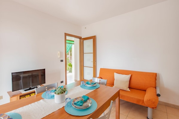 A modern living room with a wooden dining table set for two, a flat-screen TV, and a bright orange sofa.