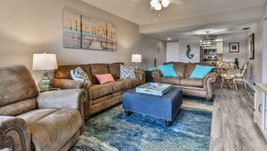 A spacious living room with two brown leather sofas, a blue ottoman, and a dining area with wicker chairs. - Castle Reef 424 Peaceful Coastal Views (New Smyrna Beach)