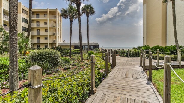 A wooden boardwalk leading to a beachfront hotel with palm trees and a clear sky. - Castle Reef 424 Peaceful Coastal Views (New Smyrna Beach)
