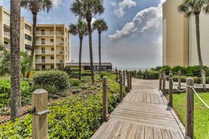 A wooden boardwalk leading to a beachfront hotel with palm trees and a clear sky. - Castle Reef 424 Peaceful Coastal Views (New Smyrna Beach)