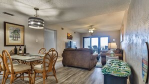 A spacious living area with a dining table, chairs, a sofa, and a TV. - Castle Reef 424 Peaceful Coastal Views (New Smyrna Beach)