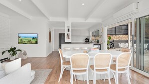 A modern kitchen with white cabinets, a dining area with a round table and wicker chairs, and a large window overlooking a garden. - Boutique Retreat Portsea by Ready Set Stay (Portsea)