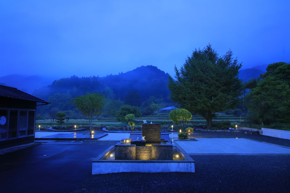 A serene outdoor area with a stone fountain, a traditional building, and a mountainous backdrop.