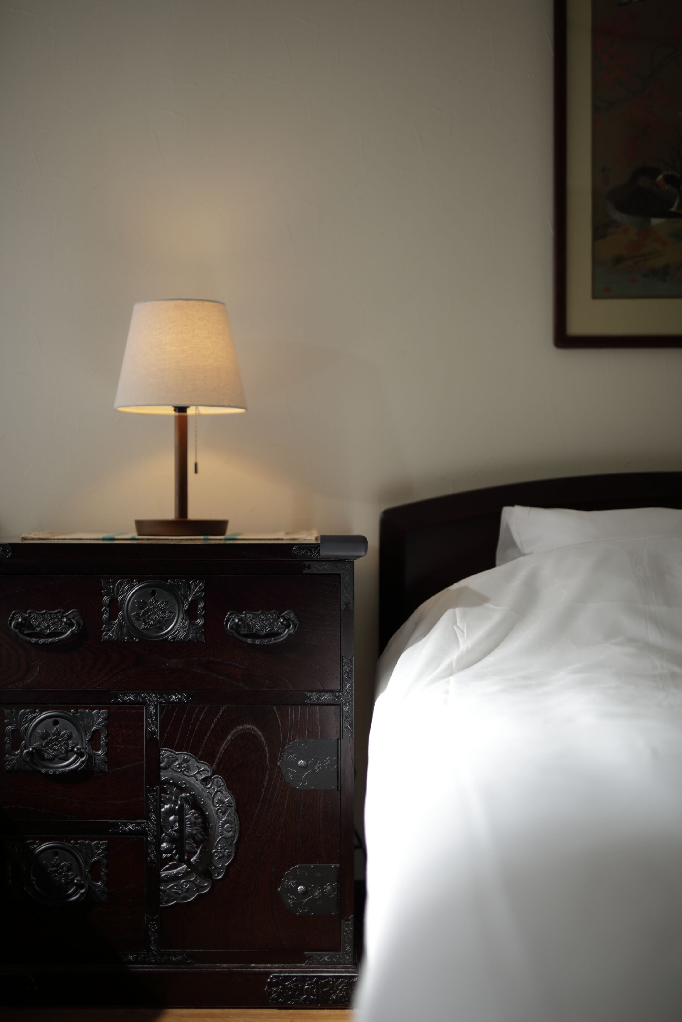 Un lit bien fait, avec du linge de lit blanc, une table de chevet en bois sur laquelle se trouve une lampe, et un tableau encadré accroché au mur.