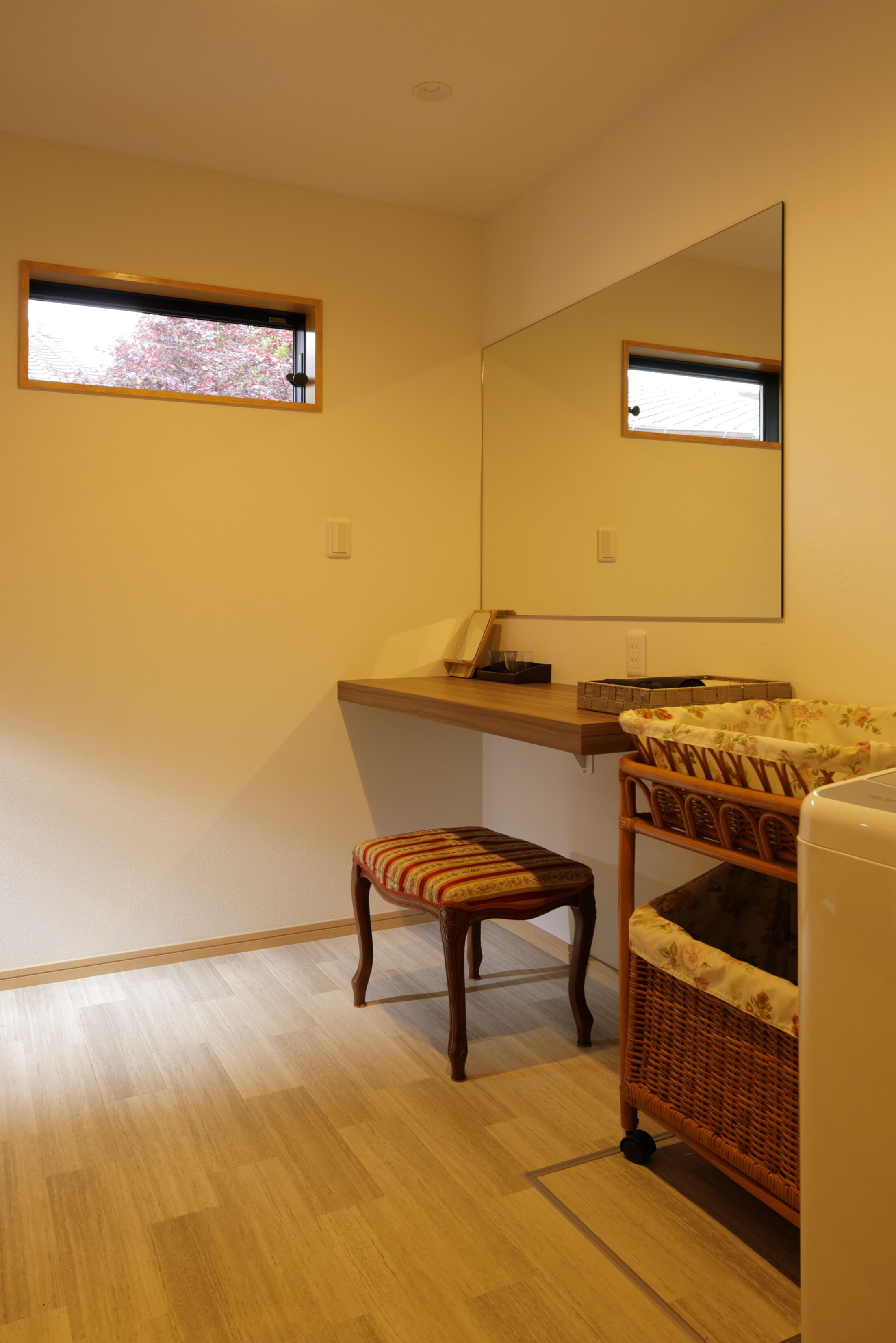 A small, well-lit room with a wooden floor, a single window, a mirror, a wicker chair, and a small stool.