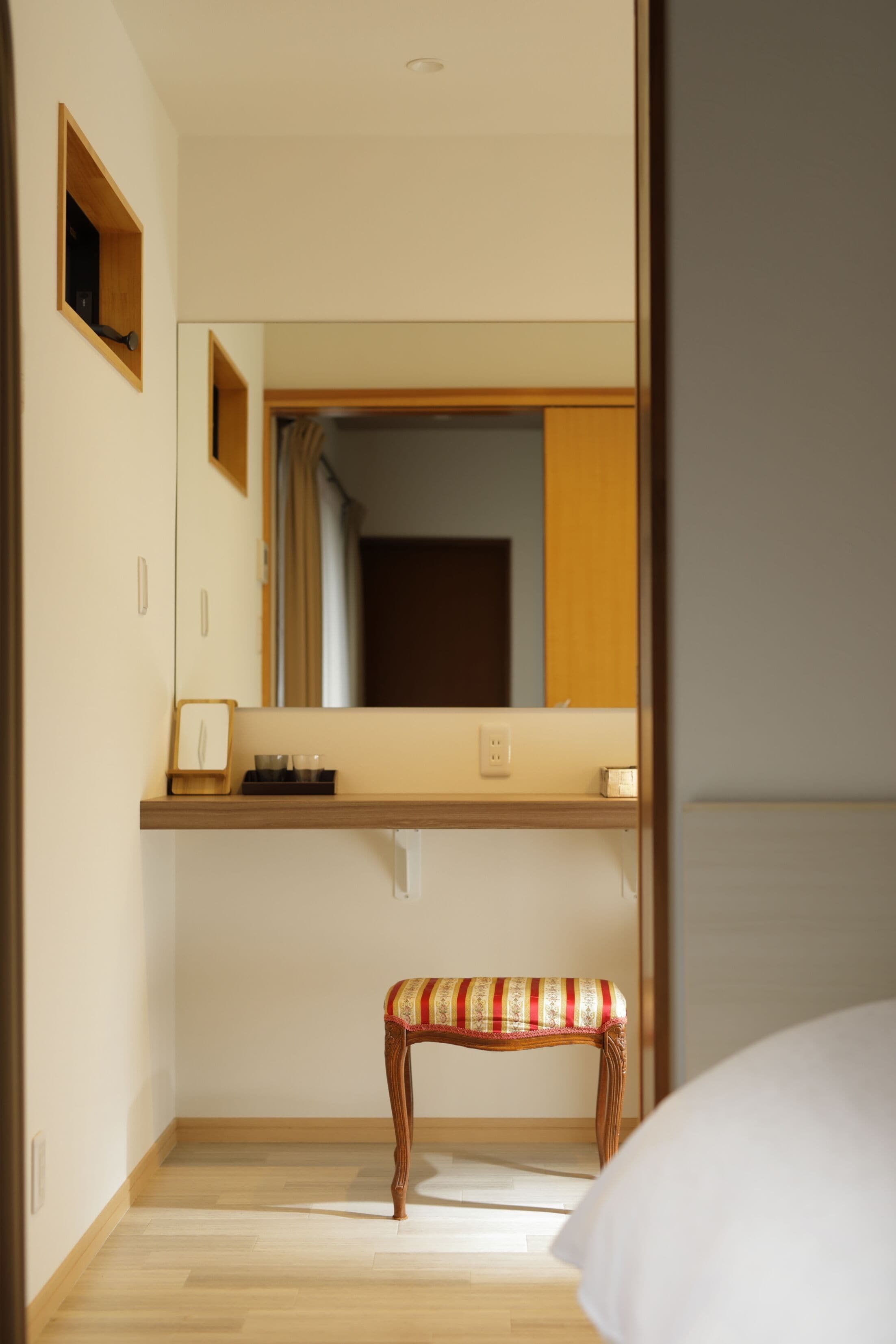Habitación de hotel con un banco de madera, una mesita de noche y un espejo que refleja una puerta y una ventana.