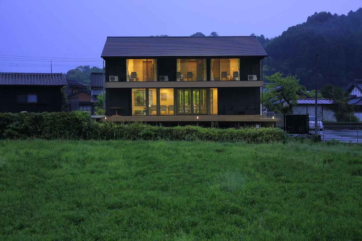 A modern two-story house with large windows and a flat roof, surrounded by greenery and hills.