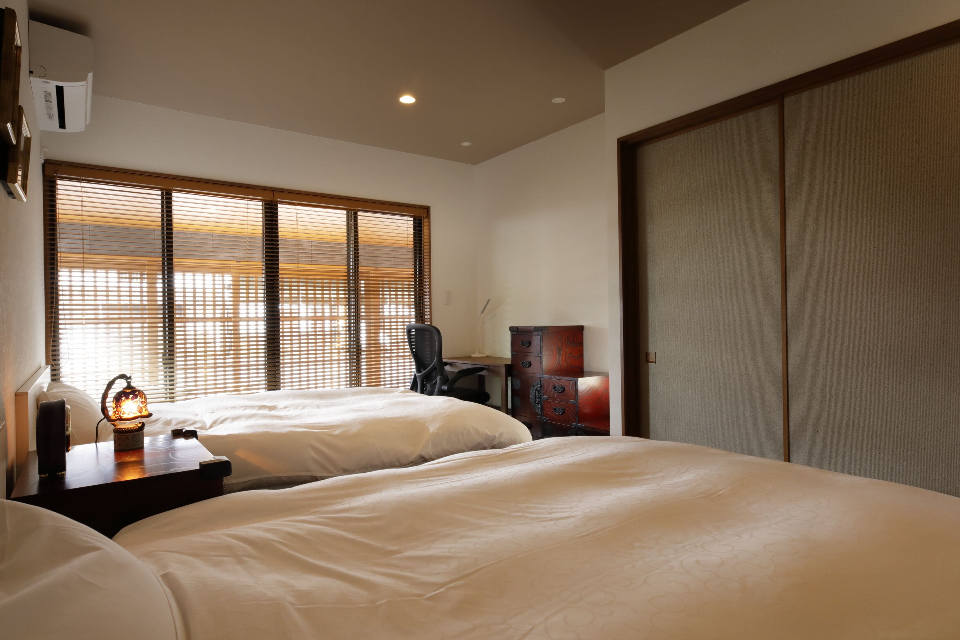 A hotel room with two beds, a desk with a chair, a wardrobe, and a large window with blinds.