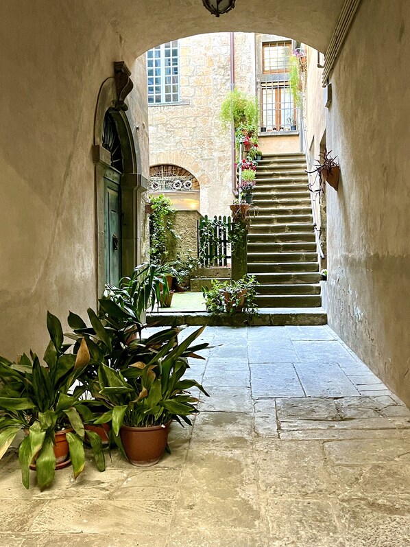 A narrow stone pathway leading to a staircase in a courtyard with potted plants and a view of a building with windows and a balcony.