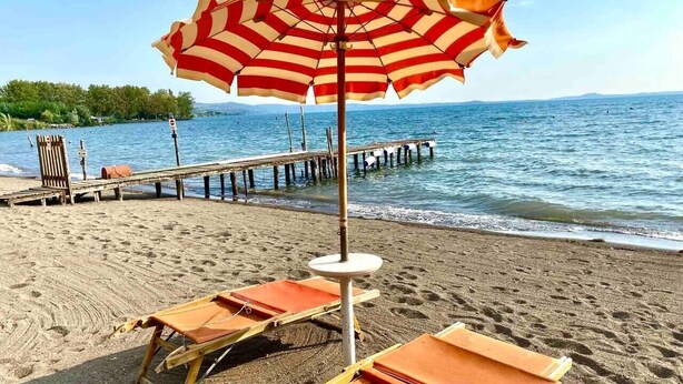 Ein Strand mit einem gestreiften Sonnenschirm, Holzliegen und einer Pier, die sich ins Wasser erstreckt.