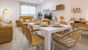 Un salon moderne comprenant un canapé, une table à manger et des chaises. Un téléviseur est fixé au mur, et une petite étagère est décorée de plantes.