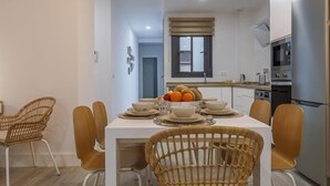 Une cuisine moderne avec des armoires blanches, un four encastré et un coin repas comprenant une table blanche et des chaises en osier.