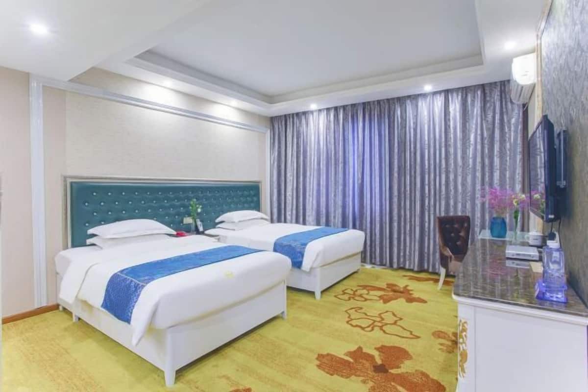 Ein Hotelzimmer mit zwei Betten, einem Schreibtisch mit Stuhl, einem Fernseher und einem großen Fenster mit Vorhängen.