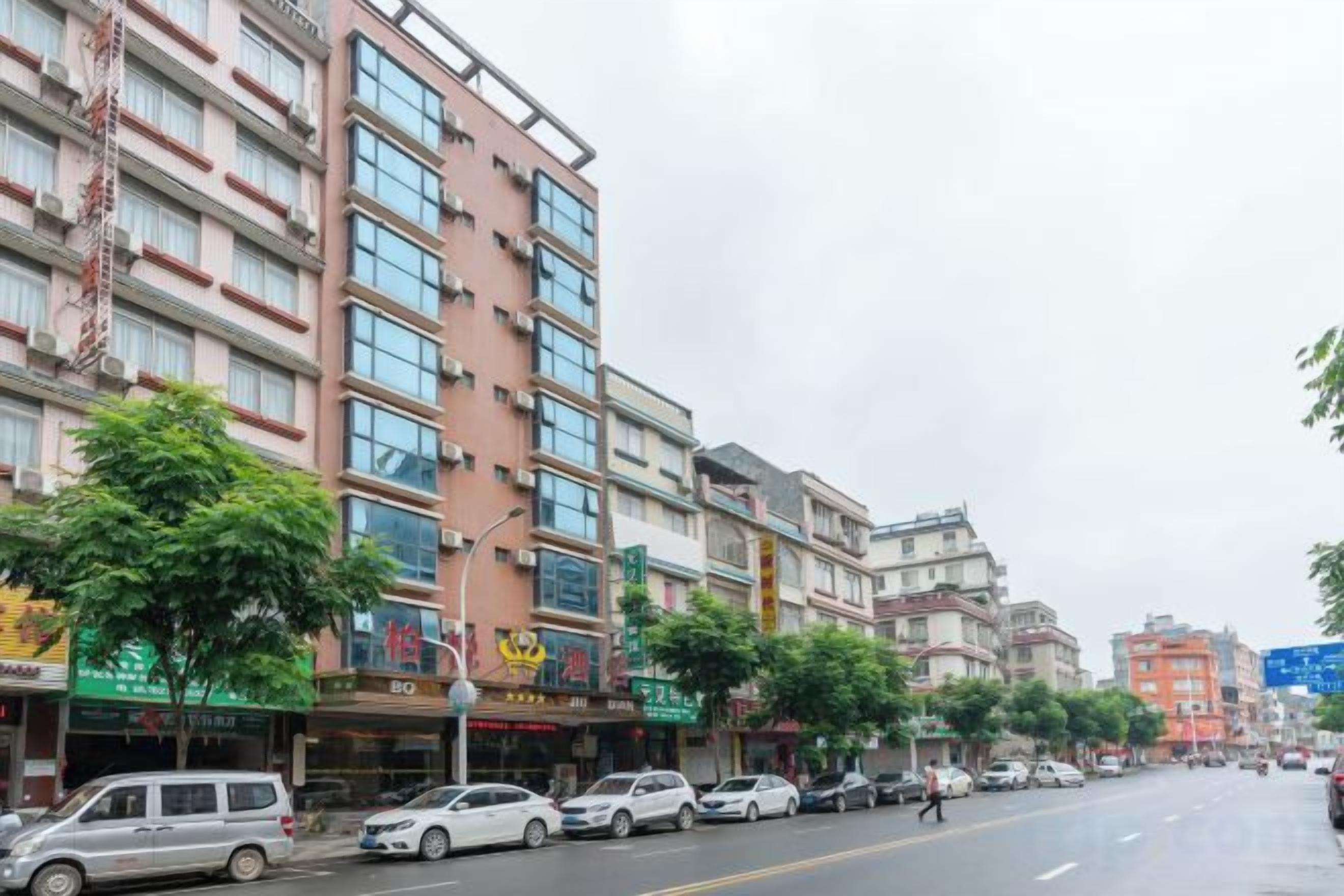 A street with multi-story buildings, parked cars, and trees.
