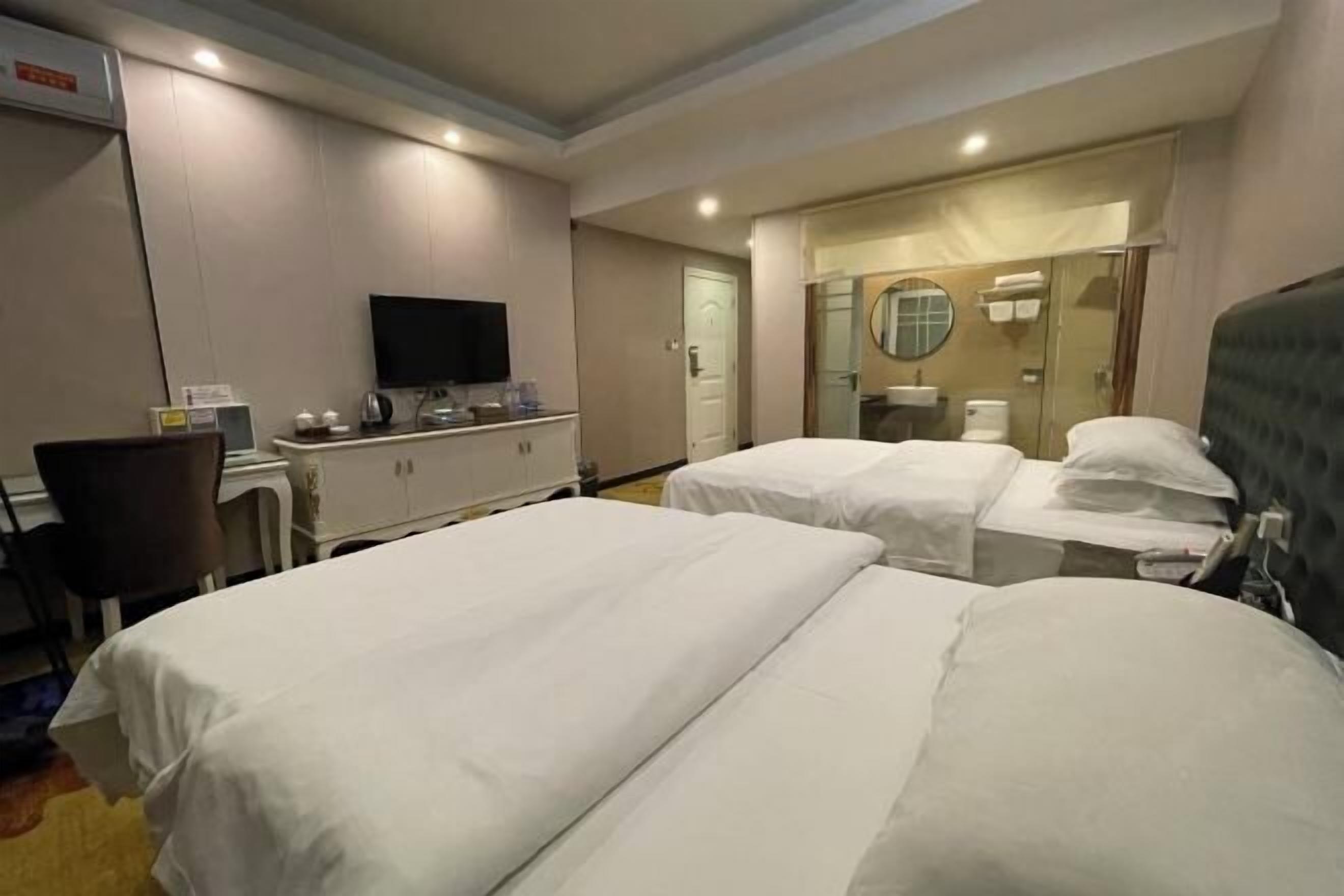 A hotel room with two beds, a TV, a desk with a chair, a mirror, and a bathroom visible through a glass door.