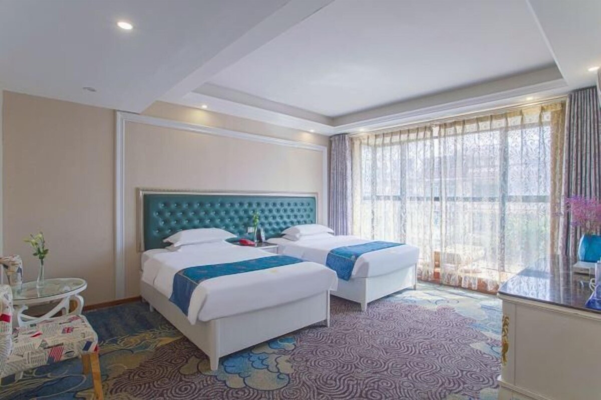 Habitación de hotel con dos camas, un ventanal con cortinas, una mesa redonda con un florero y una alfombra estampada.