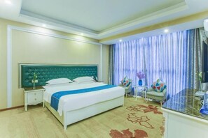 Habitación de hotel con una cama grande, una mesita de noche, una pequeña mesa con una silla, y vistas al exterior a través de cortinas.